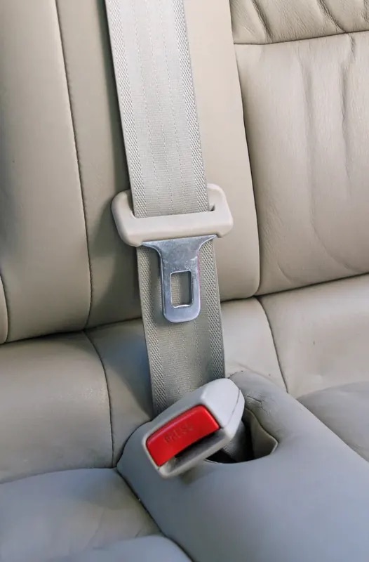 Three-point seat belt fitted on a light coloured vehicle seat, showing the shoulder webbing, latch plate and buckle